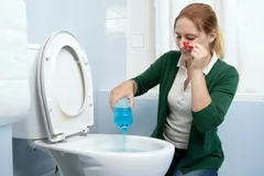 Žena čistí toaletu čisticím prostředkem a drží si nos kvůli zápachu v koupelně.