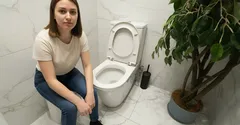 Žena sedí vedle záchodu v moderní koupelně s květinou na podlaze.