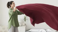 Žena ustýlá postel s vínovým přehozem v moderním ložnicovém interiéru.