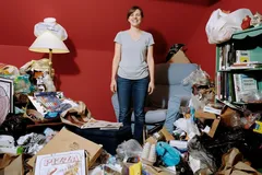 Žena stojí v místnosti plné nepořádku a hromady odpadků před červenou stěnou.